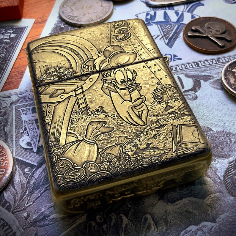 May include: A gold-colored metal lighter featuring a detailed engraved illustration of Donald Duck surrounded by money bags and coins. The design includes other cartoon characters and the number "3". The lighter is rectangular with rounded edges.