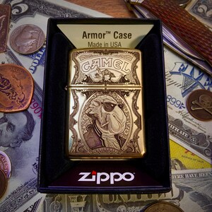 May include: A gold-colored Zippo lighter with a detailed engraving of a camel wearing sunglasses, labeled "Joe Camel." The lighter is in a black box with the "Armor Case" and "Made in USA" text. The Zippo logo is visible.
