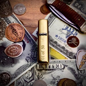 May include: A gold-colored lighter with the text "01 of 50" is displayed on a wooden surface. The lighter is surrounded by vintage currency, coins, and a pocket knife. The scene evokes a sense of history and collecting.