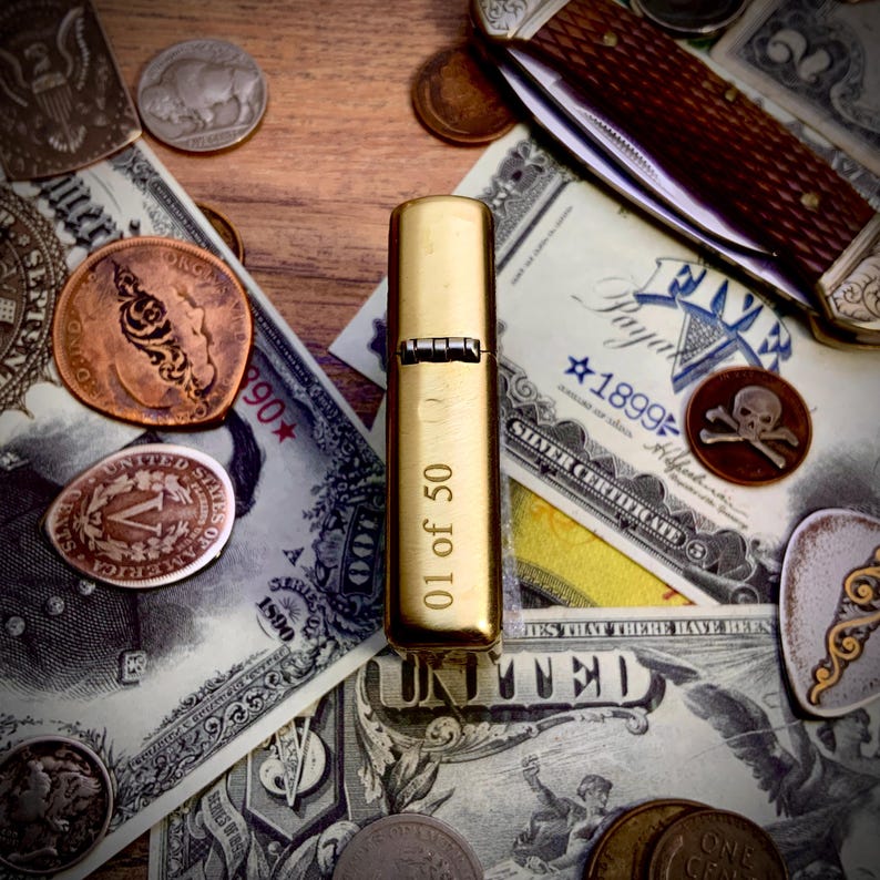May include: A gold-colored lighter with the text "01 of 50" is displayed on a wooden surface. The lighter is surrounded by vintage currency, coins, and a pocket knife. The scene evokes a sense of history and collecting.