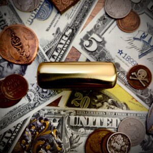 May include: A gold-colored, rectangular object rests atop a collection of vintage currency, including dollar bills, coins, and a copper penny. The bills display text such as "United States" and "Five Dollars."