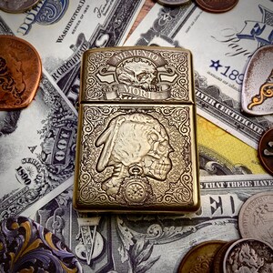 May include: A brass-colored lighter with intricate engraved designs. The top features a skull and the words "Memento Mori." The bottom half depicts a Native American profile with a skull-like appearance. The lighter is surrounded by currency and coins.