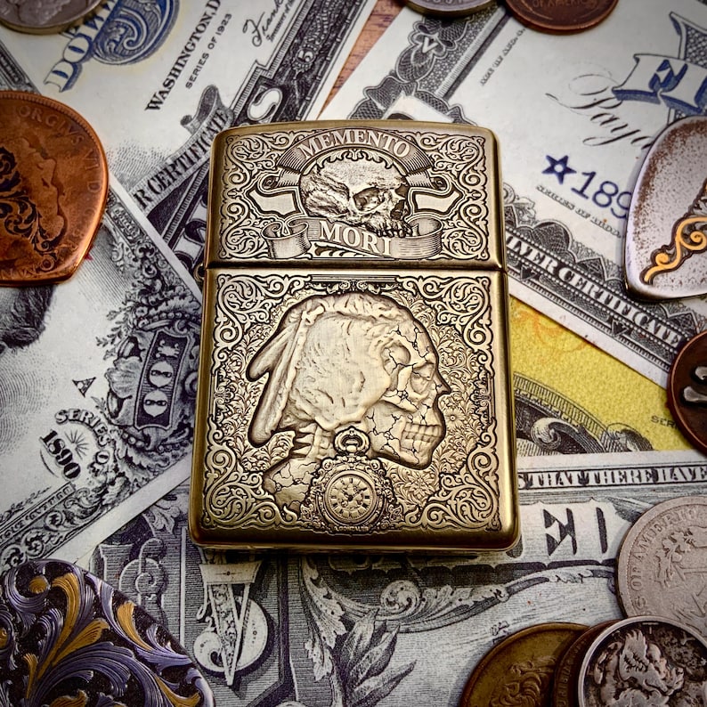 May include: A brass-colored lighter with intricate engraved designs. The top features a skull and the words "Memento Mori." The bottom half depicts a Native American profile with a skull-like appearance. The lighter is surrounded by currency and coins.