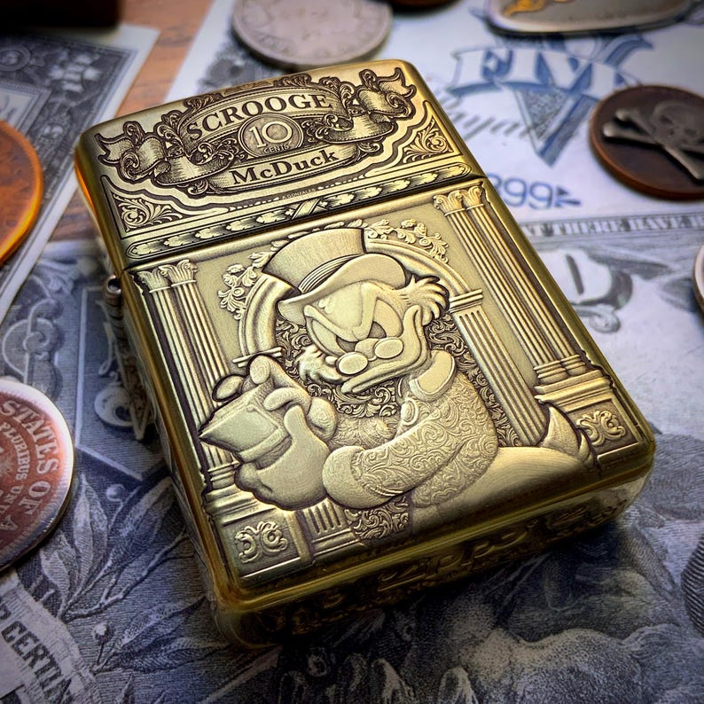 May include: A gold-colored metal lighter featuring an engraved image of Scrooge McDuck holding a money bag. The lighter has the text "SCROOGE McDuck" and "10 SERIES" engraved on the top.