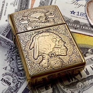May include: A gold-colored metal lighter with intricate engraved designs. The top features a skull and the words "MEMENTO MORI." The front displays a detailed Native American skull profile. The lighter is rectangular and has a decorative border.