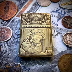 May include: A gold-colored metal lighter featuring an embossed image of Scrooge McDuck holding a money bag. The lighter has the text "SCROOGE McDuck" and is surrounded by coins and dollar bills.