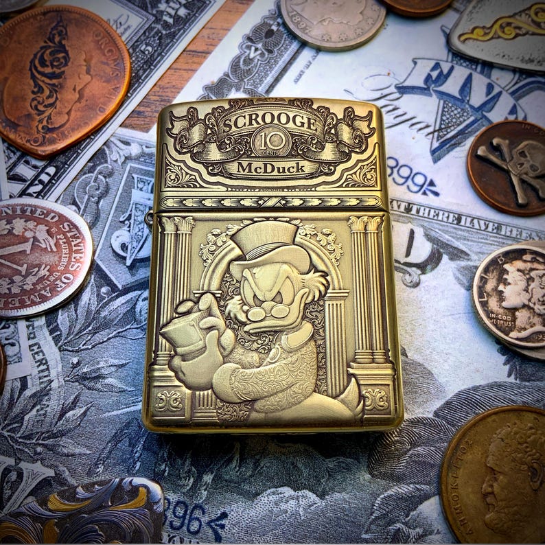 May include: A gold-colored metal lighter featuring an embossed image of Scrooge McDuck holding a money bag. The lighter has the text "SCROOGE McDuck" and is surrounded by coins and dollar bills.