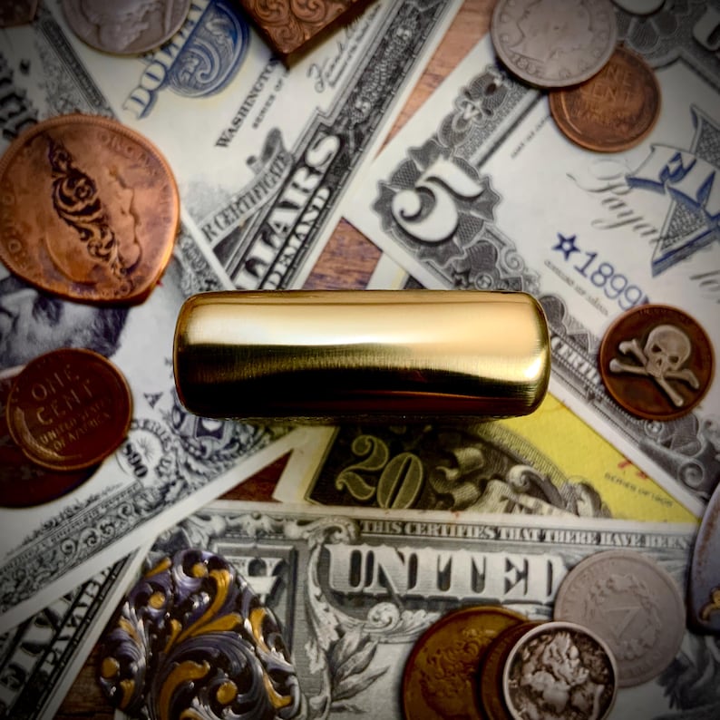 May include: A gold-colored, rectangular object rests atop a collection of vintage currency, including dollar bills, coins, and a copper penny. The bills display text such as "United States" and "Five Dollars."