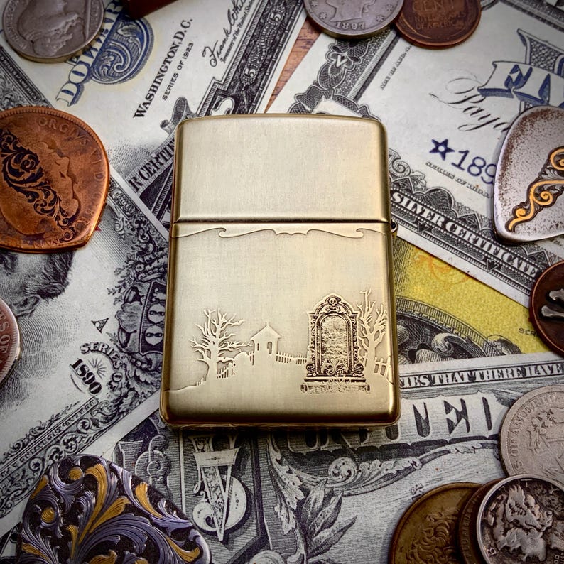May include: A gold-colored lighter with an engraved graveyard scene. The design includes a tombstone, trees, and a small building. The lighter is rectangular and has a hinged lid.
