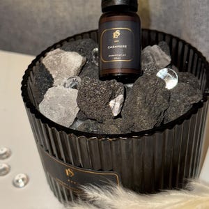 Può includere: Un contenitore di vetro nero pieno di rocce grigio scuro e cristalli trasparenti, che contiene una piccola bottiglia di olio essenziale marrone con l'etichetta "Cashmere". La bottiglia ha un tappo nero e un'etichetta dorata.