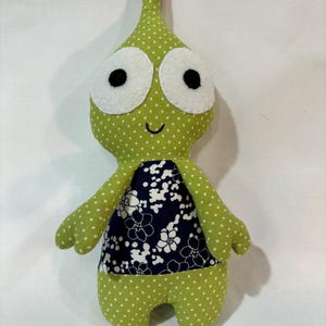 Puede incluir: Un juguete de peluche verde con lunares blancos, ojos grandes blancos con pupilas negras y un sombrero rojo y blanco. El juguete tiene un patrón floral azul y blanco en su torso.
