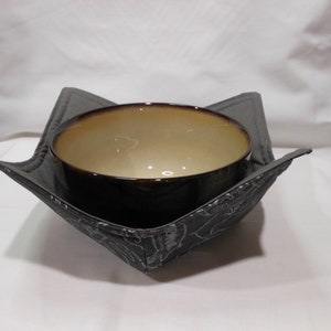 Puede incluir: Un cuenco redondo de cerámica con exterior marrón oscuro e interior beige claro, colocado dentro de un soporte de tela cuadrado. El soporte está hecho de tela acolchada gris oscuro con un sutil patrón de líneas blancas de perros.