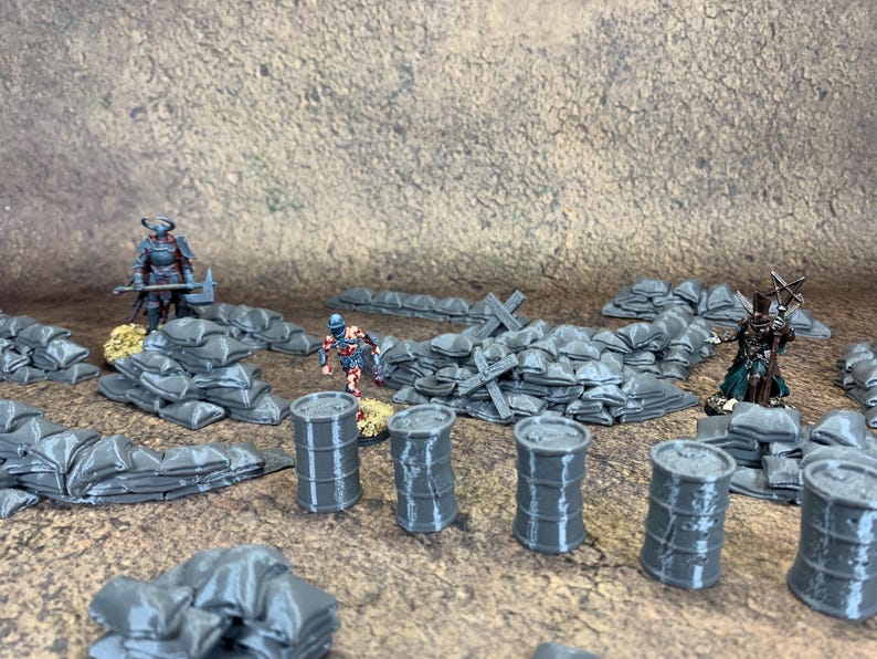 May include: A miniature war scene featuring gray sandbags, barrels, and figures on a textured surface. The scene includes several small figures, sandbags, and barrels. The figures are painted with detailed armor and are set in a war-like environment.