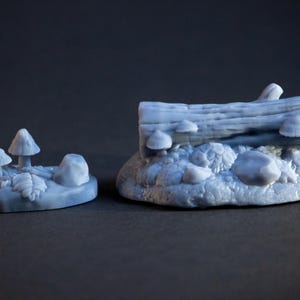 Könnte beinhalten: Zwei Miniatur-Waldszenen mit Pilzen und einem Baumstamm. Die erste Szene zeigt eine Gruppe von drei Pilzen auf einer Basis mit einem kleinen Stein. Die zweite Szene zeigt einen Baumstamm mit Pilzen, die darauf wachsen, umgeben von Steinen und Blättern.
