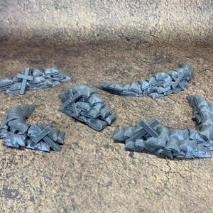 May include: Five grey miniature barricades, each made of sandbags and wooden crosses. The sandbags are stacked to form a wall, and the crosses are placed on top. The pieces are designed for tabletop gaming.