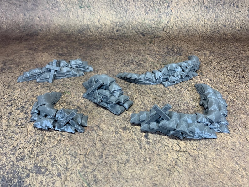 May include: Five grey miniature barricades, each made of sandbags and wooden crosses. The sandbags are stacked to form a wall, and the crosses are placed on top. The pieces are designed for tabletop gaming.
