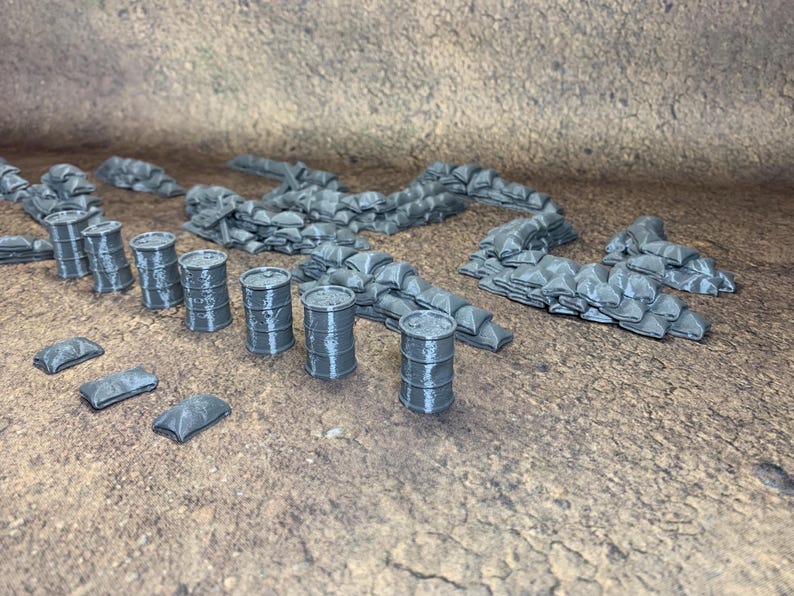 May include: A collection of grey miniature barrels, sandbags, and barricades. These 3D-printed models are designed for tabletop gaming or model displays. The set includes several barrels, sandbag walls, and small barricades, all in a neutral grey colour.