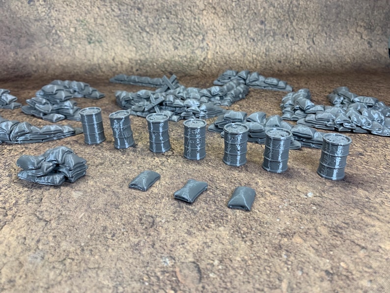 May include: Miniature grey terrain pieces for tabletop gaming, including sandbags, barrels, and small rectangular objects. The pieces are arranged on a textured brown surface. These items are designed for miniature wargaming.