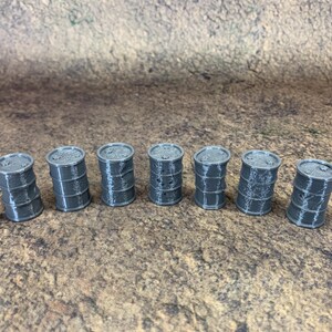 May include: Seven small, grey barrels are arranged in a row. Each barrel has a cylindrical shape with horizontal bands and a flat top. They are set on a textured, brown surface. Each barrel is approximately 3 cm tall.