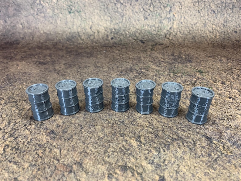 May include: Seven small, grey barrels are arranged in a row. Each barrel has a cylindrical shape with horizontal bands and a flat top. They are set on a textured, brown surface. Each barrel is approximately 3 cm tall.