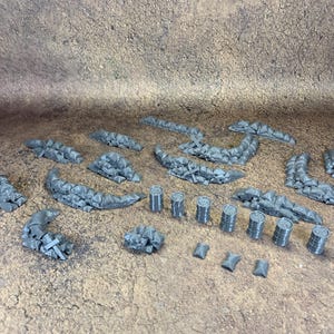 May include: A selection of grey miniature terrain pieces, including sandbag walls, barrels, and barricades. The pieces are arranged on a textured brown surface, ideal for tabletop gaming or model building.