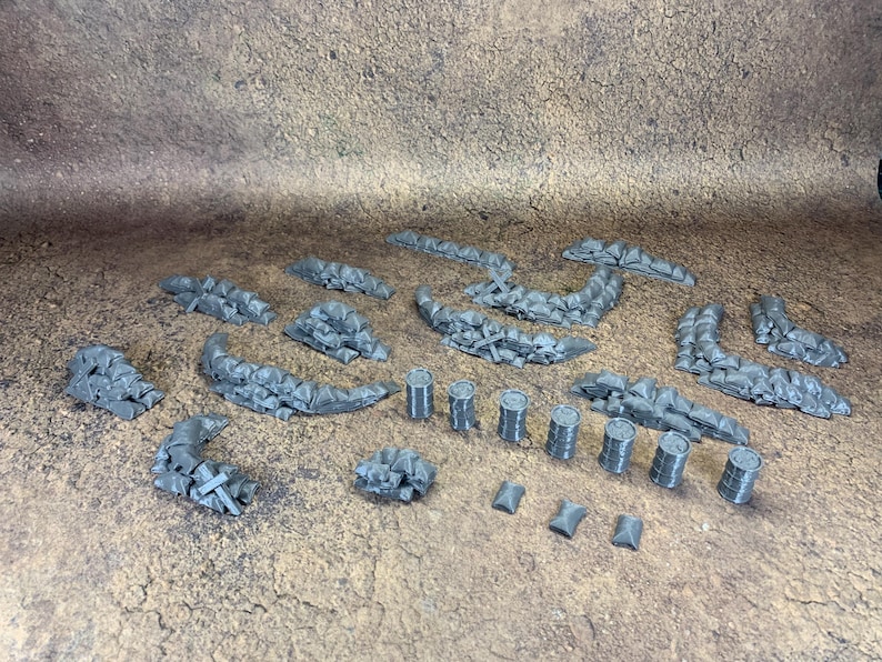 May include: A selection of grey miniature terrain pieces, including sandbag walls, barrels, and barricades. The pieces are arranged on a textured brown surface, ideal for tabletop gaming or model building.