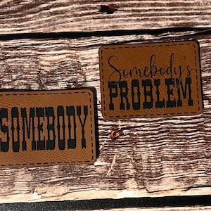 May include: Two brown leather patches with black text. One patch reads "SOMEBODY" and the other reads "Somebody's PROBLEM". Both patches have a stitched border and are set against a wooden background.