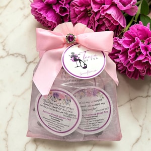 May include: A pink gift bag tied with a pink ribbon and a decorative jewel. The bag contains two tea sachets with text and floral designs. The background features vibrant pink flowers.