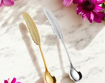 May include: Two metal spoons with handles shaped like detailed feathers. One spoon has a polished gold finish, and the other has a polished silver finish. Both spoons feature a long, slender stem and a rounded bowl at the end of the handle.