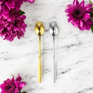 May include: Two spoons with rose-shaped bowls. One spoon is gold-colored, the other is silver. The spoons are on a marble surface, with pink flowers in the background. The spoons have long, slender handles.