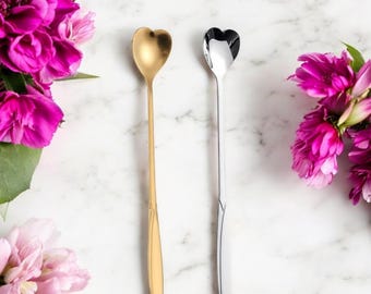 May include: Two heart-shaped spoons, one gold and one silver, are displayed against a marble background. The spoons have long, slender handles and are positioned vertically. Pink flowers are arranged on either side of the spoons.