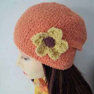 Op de afbeelding: Een gebreide beanie in een perzikkleurige oranje met een gele en bruine bloemversiering.