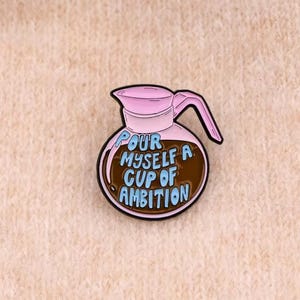Peut inclure: Une broche en émail métallique en forme de cafetière avec un couvercle et une anse rose clair. Le corps de la cafetière est rempli d'un liquide brun foncé et porte le texte bleu clair "POUR MYSELF A CUP OF AMBITION" en lettres arrondies.