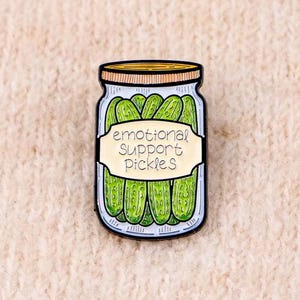 May include: A metal enamel pin shaped like a glass jar filled with green pickles. The jar features a tan label with the black text 'emotional SUPPORT pickles' and has a tan lid. The design is outlined in black with a white background.