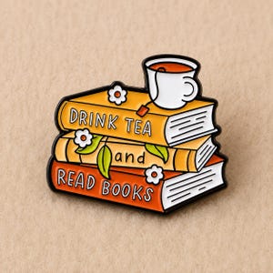 Peut inclure: Une broche en émail métallique représentant une pile de trois livres aux couvertures orange et jaune. Le livre du haut porte l'inscription DRINK TEA, le milieu and, et le bas READ BOOKS. Une tasse blanche avec un sachet de thé et des fleurs.