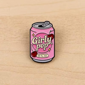 May include: A pink enamel pin shaped like a soda can with the text Girly pop in a cream-coloured font. The design features two red cherries with stems and small yellow star accents. The bottom of the can displays the text 12 FL. OZ. in black.