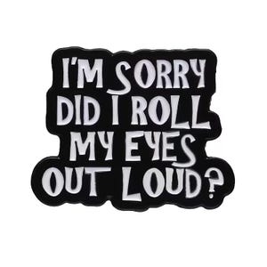 Peut inclure: Une broche en émail noir avec des lettres blanches qui dit, I'M SORRY DID I ROLL MY EYES OUT LOUD? Le texte est disposé sur quatre lignes avec une police grasse et stylisée, et la broche présente une bordure noire suivant le contour.
