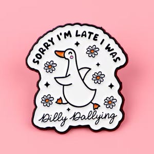 Peut inclure: Une broche en émail blanc représentant un canard de dessin animé avec un bec et des pattes orange, entouré de petites fleurs blanches et d'étoiles noires. Le texte SORRY I'M LATE I WAS DILLY DALLYING est écrit en noir sur fond blanc.