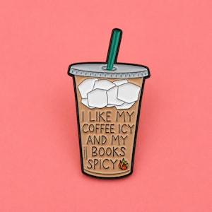 May include: A tan enamel pin shaped like a coffee cup with a lid and a green straw. The cup contains white ice cubes and features the black text I LIKE MY COFFEE ICY AND MY BOOKS SPICY with a small orange and red flame icon at the bottom.