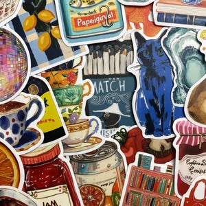 Peut inclure: Un assortiment d'autocollants colorés avec divers motifs. Les autocollants représentent un chat bleu, une canette de jus d'orange, un pot de confiture, une étagère, des citrons, des tasses à thé et une boîte de sardines avec le texte "Paperback".