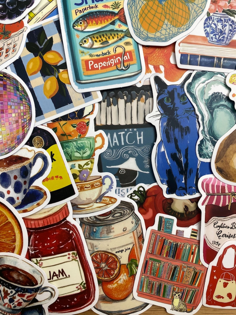 Op de afbeelding: Een verzameling kleurrijke stickers met diverse ontwerpen. Stickers tonen een blauwe kat, een blikje sinaasappelsap, een pot jam, een boekenplank, citroenen, theekopjes en een sardineblikje met de tekst "Paperback".