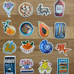 Op de afbeelding: Een verzameling stickers met verschillende illustraties. Inbegrepen zijn een pizza, een konijn, een ketchupfles, een kom ontbijtgranen, een papaja, een blikje sardines, een goudvis, een kikker en een doosje lucifers.