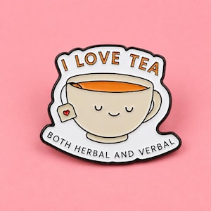 May include: A white enamel pin shaped like a tea cup with a smiling face, filled with orange liquid and a tea bag tag featuring a red heart. The text reads I LOVE TEA at the top and BOTH HERBAL AND VERBAL at the bottom in black lettering.