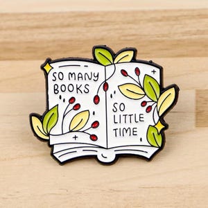 Peut inclure: Une broche en émail métallique en forme de livre ouvert avec des pages blanches et des contours noirs. Le texte indique SO MANY BOOKS SO LITTLE TIME. Le motif présente des feuilles vertes et jaunes avec de petites baies rouges et des étoiles jaunes.