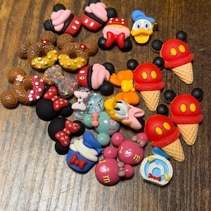 May include: A collection of small, colorful resin charms featuring various cartoon-inspired designs, including Mickey Mouse-shaped donuts, ice cream cones, bows, and character faces in shades of red, pink, yellow, blue, and brown.