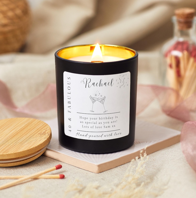 May include: Black candle with a gold lid and a white label that says "40 & FABULOUS Rachael Hope your birthday is as special as you are! Lots of love Sam xx Hand-poured with love" with a graphic of two champagne glasses.
