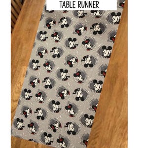 May include: Gray Mickey Mouse table runner. The fabric features a repeating pattern of Mickey Mouse faces in a circular design with a starburst effect. The text "MICKEY TABLE RUNNER" is displayed in a white rectangle.