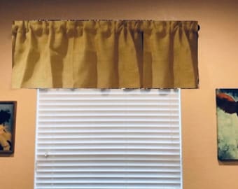 May include: A tan valance hangs above white horizontal blinds. The valance is gathered at the top, creating soft folds. Two framed cow paintings flank the window, adding a rustic touch to the room's decor.