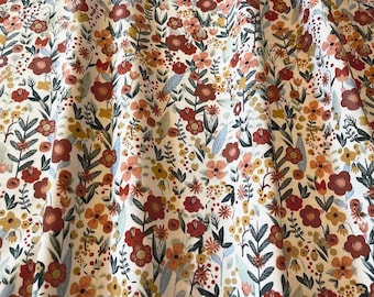 May include: A white fabric featuring a dense, repeating pattern of wildflowers in shades of rust red, mustard yellow, soft pink, and muted sage green. The design includes various flower shapes, thin stems, and small leaves arranged in a vertical flow.