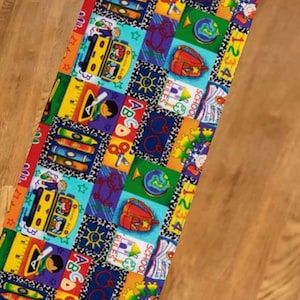 May include: A colorful fabric table runner with a school theme. The runner features a repeating pattern of school buses, books, pencils, apples, globes, and the letters of the alphabet. The background is a patchwork of bright colors, including red, yellow, blue, green, and orange.
